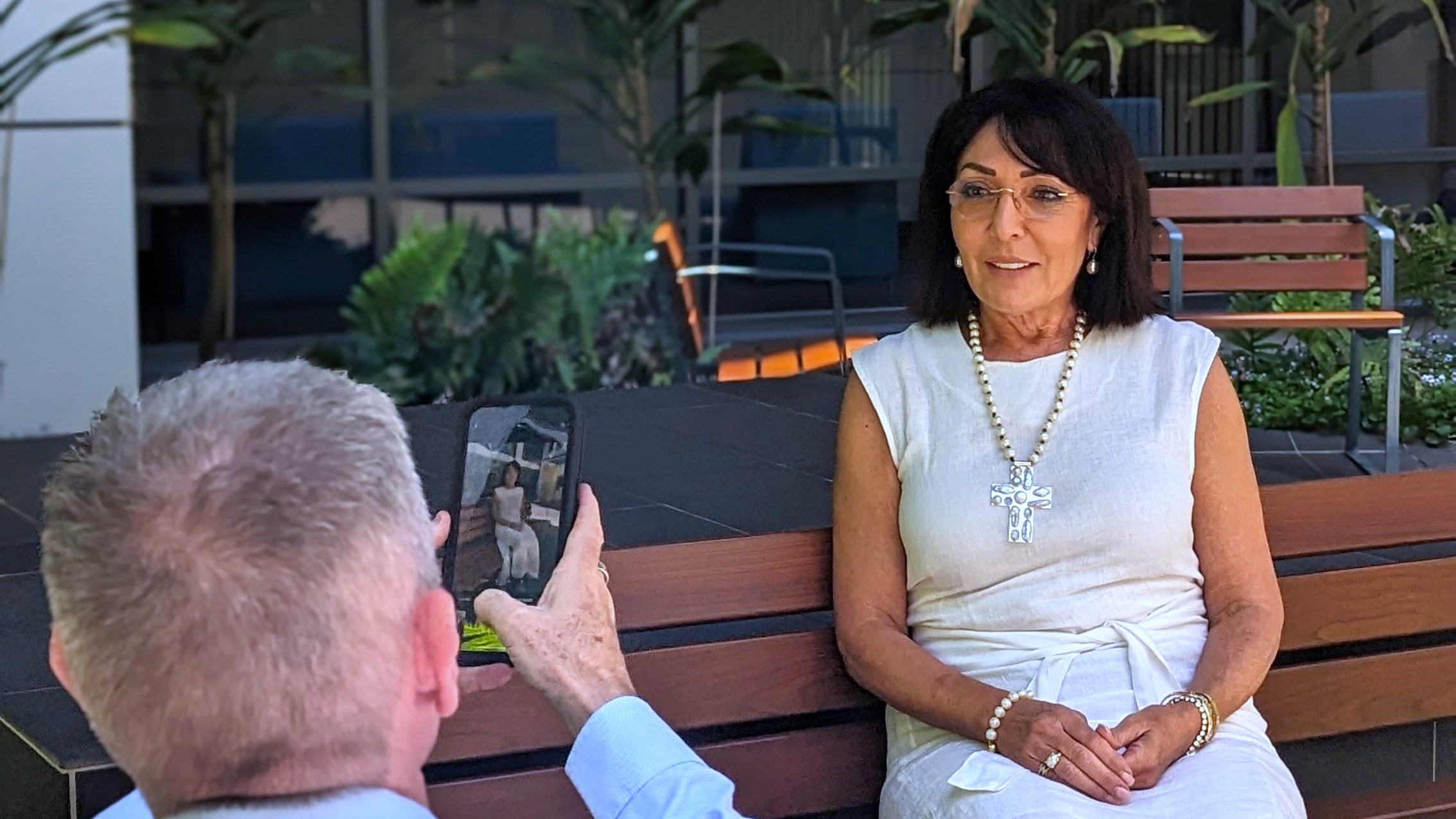 A woman sits on a bench having her photo taken by a man, who we see the back of as he holds up his iPhone to snap the picture.