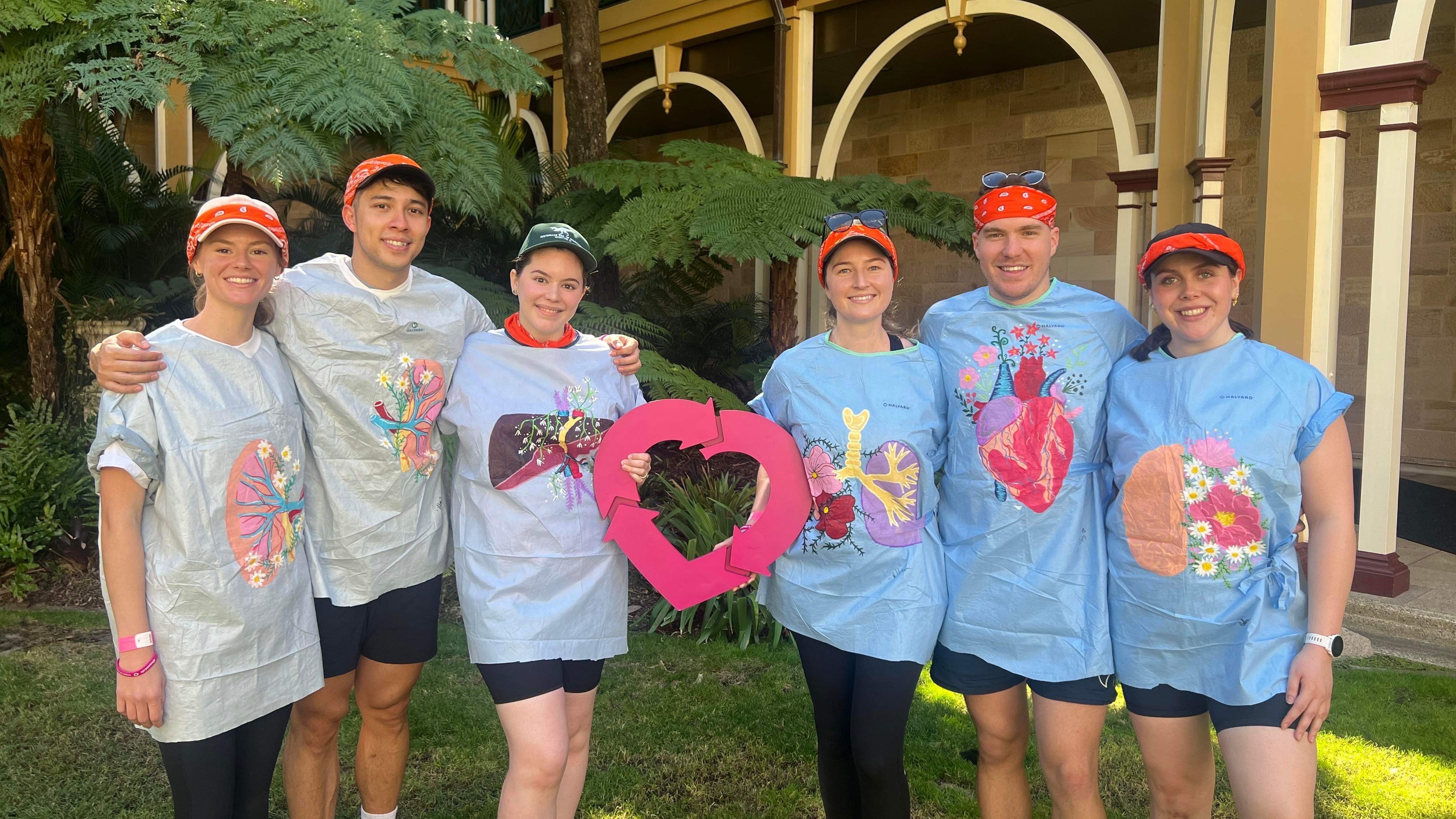 Six people pose holding a DonateLife heart logo. They are all wearing surgical gowns that have artworks of organs and flowers painted on the front of them.  