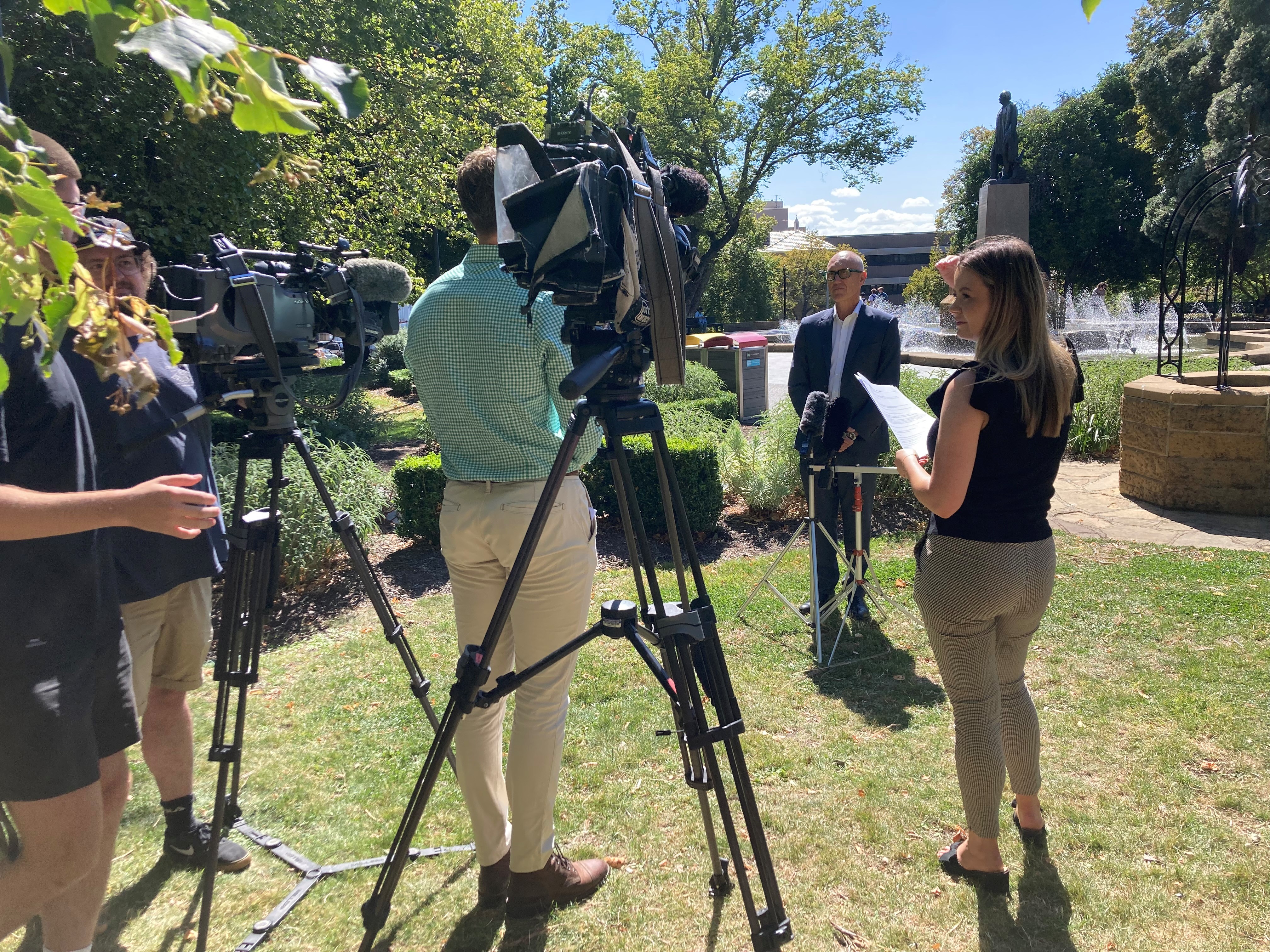 A man stands outside in a garden, speaking in front of media reporters with cameras and microphones.