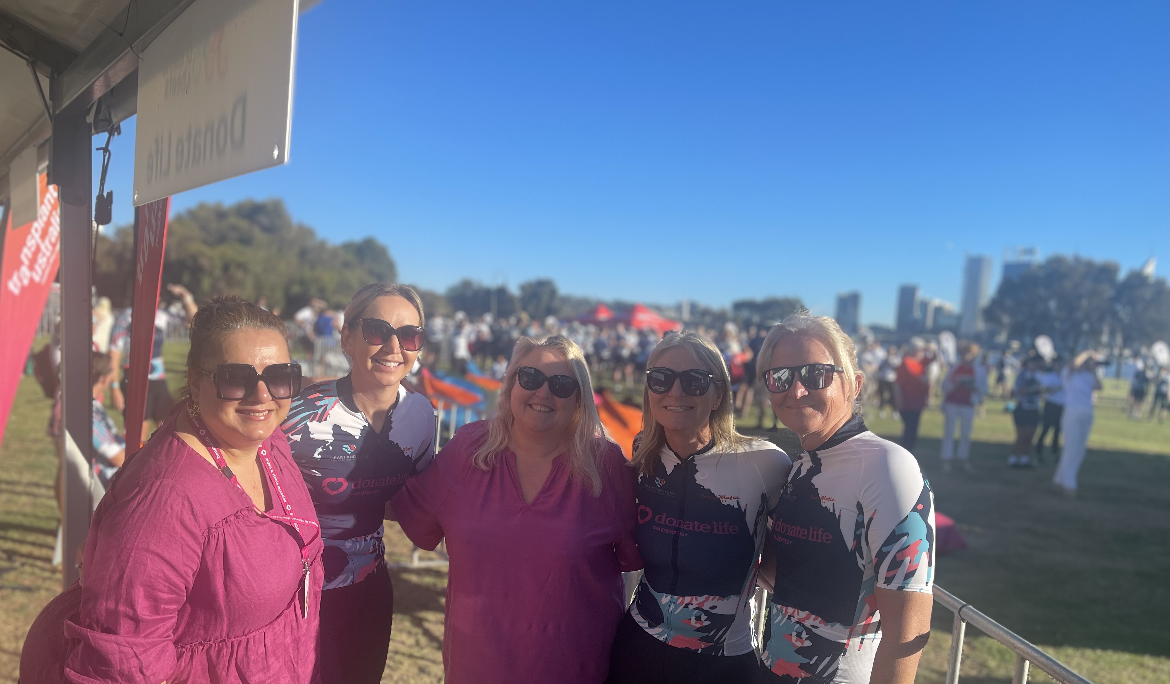 DonateLife staff pose for a photo: three of them are dressed in riding gear. They are outside with a blue sky and city buildings in the background.