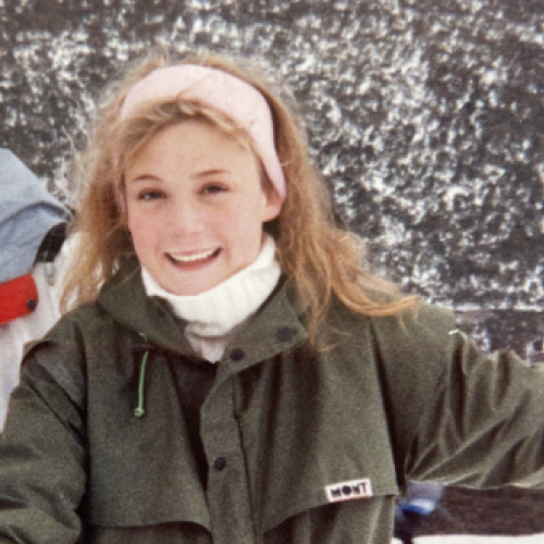 woman standing in front of a snowy mountain. She's wearing a coat zipped up to her chin, a pink headband and is holding skiing poles.