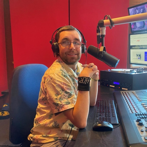 Man in a red radio studio, wearing headphones and glasses, surrounded by sound equipment, smiling at the camera.